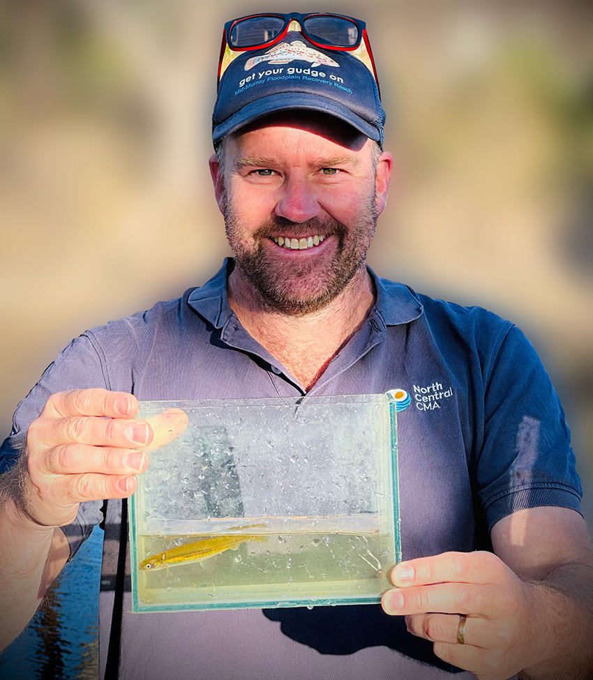 Man with a beard and wearing a cap holding a small fish tank with small-bodies fish in it.